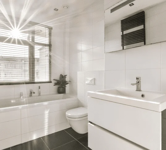 Bright, sunlit bathroom with white tiles, modern fixtures, and a plant accent. Average Cost of Small Bathroom Remodel.