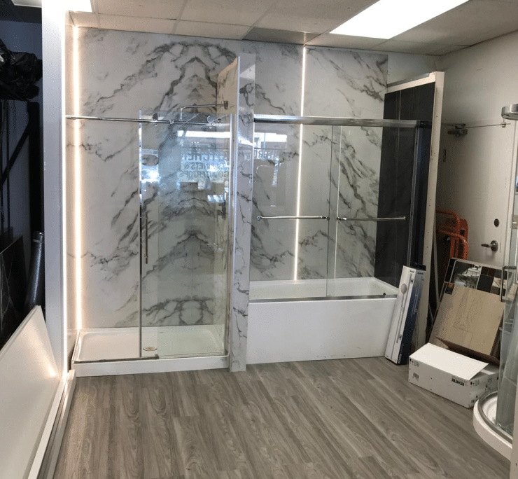 Inside a bath and kitchen showroom featuring a mockup of a marble-tiled shower area with glass doors and LED-lit mirrors, paired with gray wood-patterned flooring.