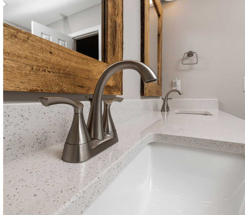 Modern bathroom faucet with a brushed finish on a speckled countertop, complemented by a rustic wooden mirror frame, in a bathroom showroom setting.