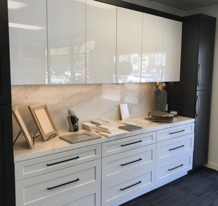 An array of sleek white cabinetry with modern black handles on display in a cabinet showroom, accented by a stone backsplash.