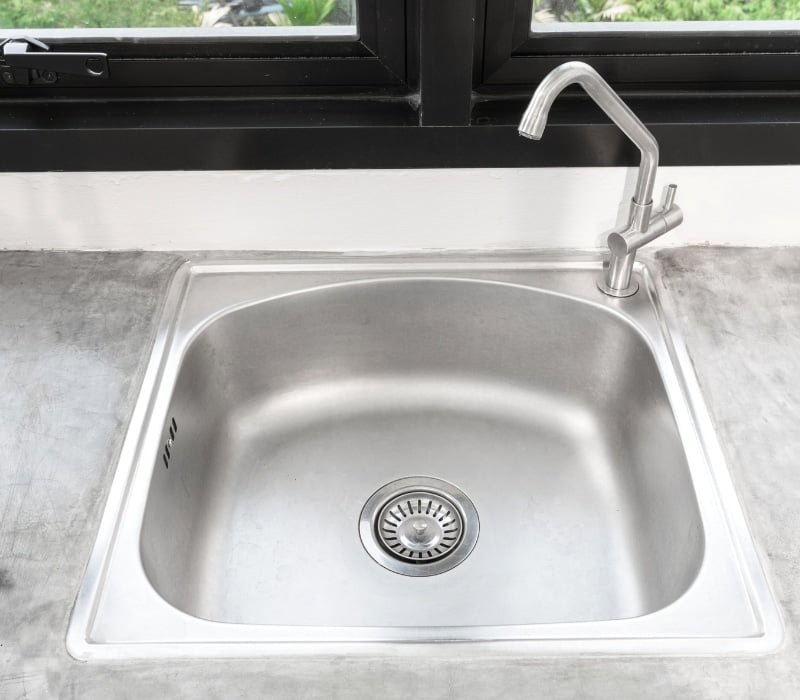 Close-up of a stainless-steel sink with a modern faucet set against a concrete countertop, featuring a garden view through the window above