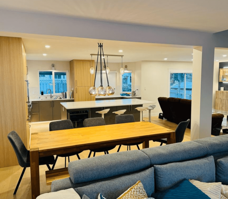 Open-concept kitchen with wooden cabinets, white island, hanging lights, and a wooden dining table adjacent to a living area.