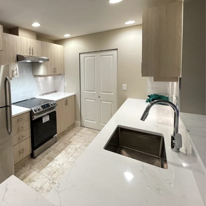 Compact kitchen with light wood cabinets, white quartz countertops, stainless steel appliances, and tiled flooring.