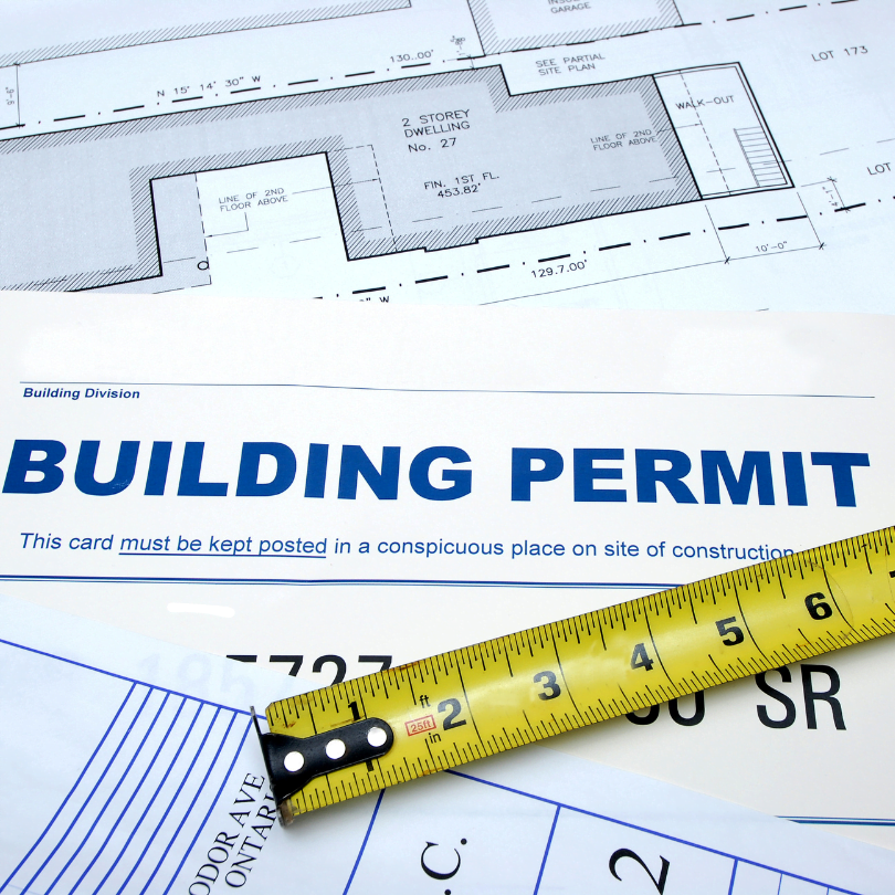 A building permit and architectural plans, with a yellow measuring tape placed across them.